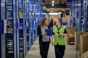 Two women looking at each other and smiling while walking through a warehouse holding clipboards.